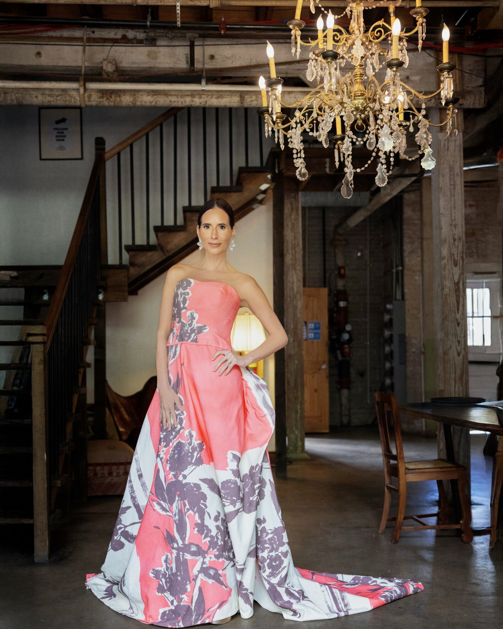 Woman in a pink and purple floral gown standing indoors near a staircase and chandelier.