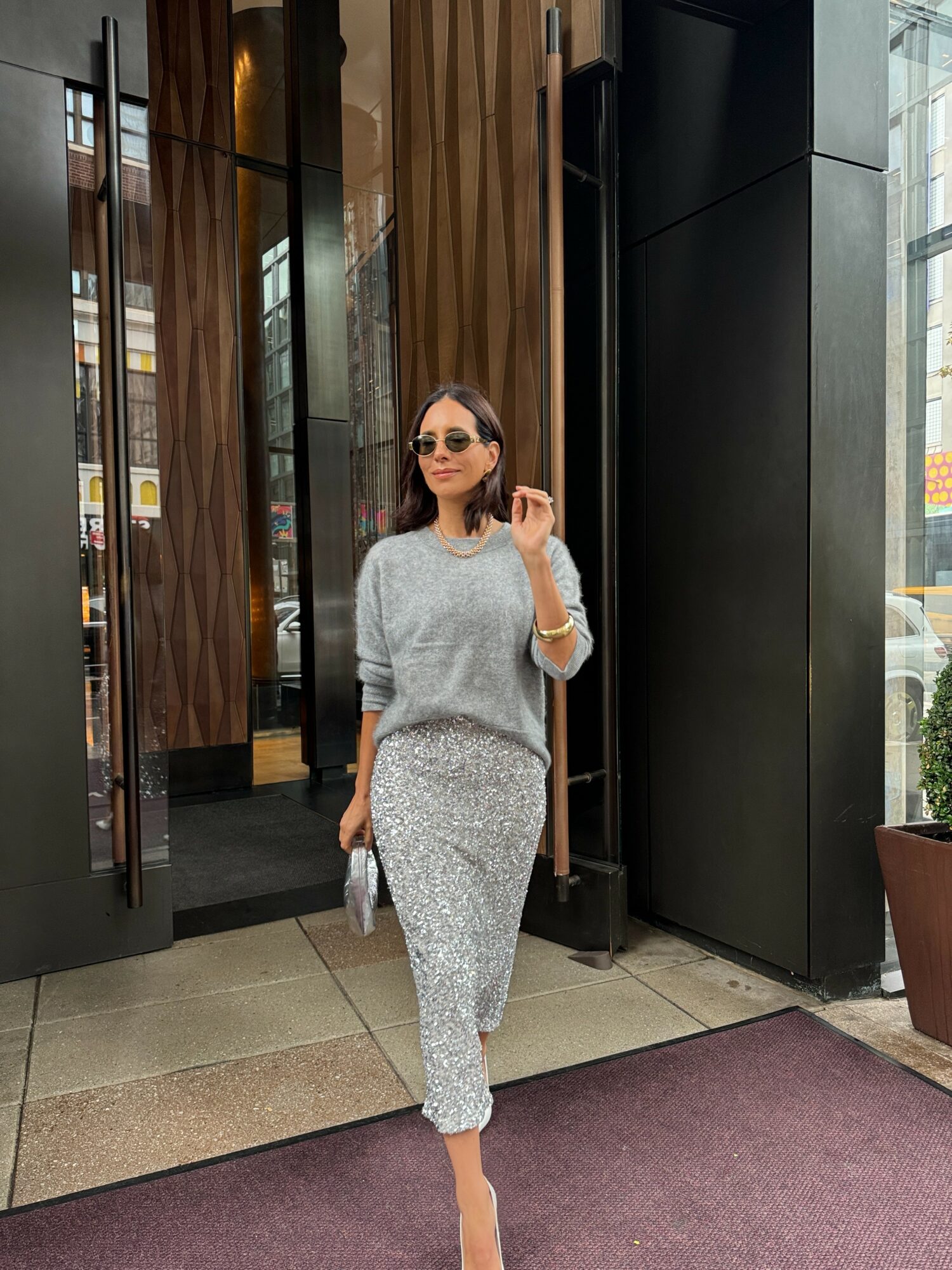 Woman in sunglasses and light-colored outfit walking outside building entrance.