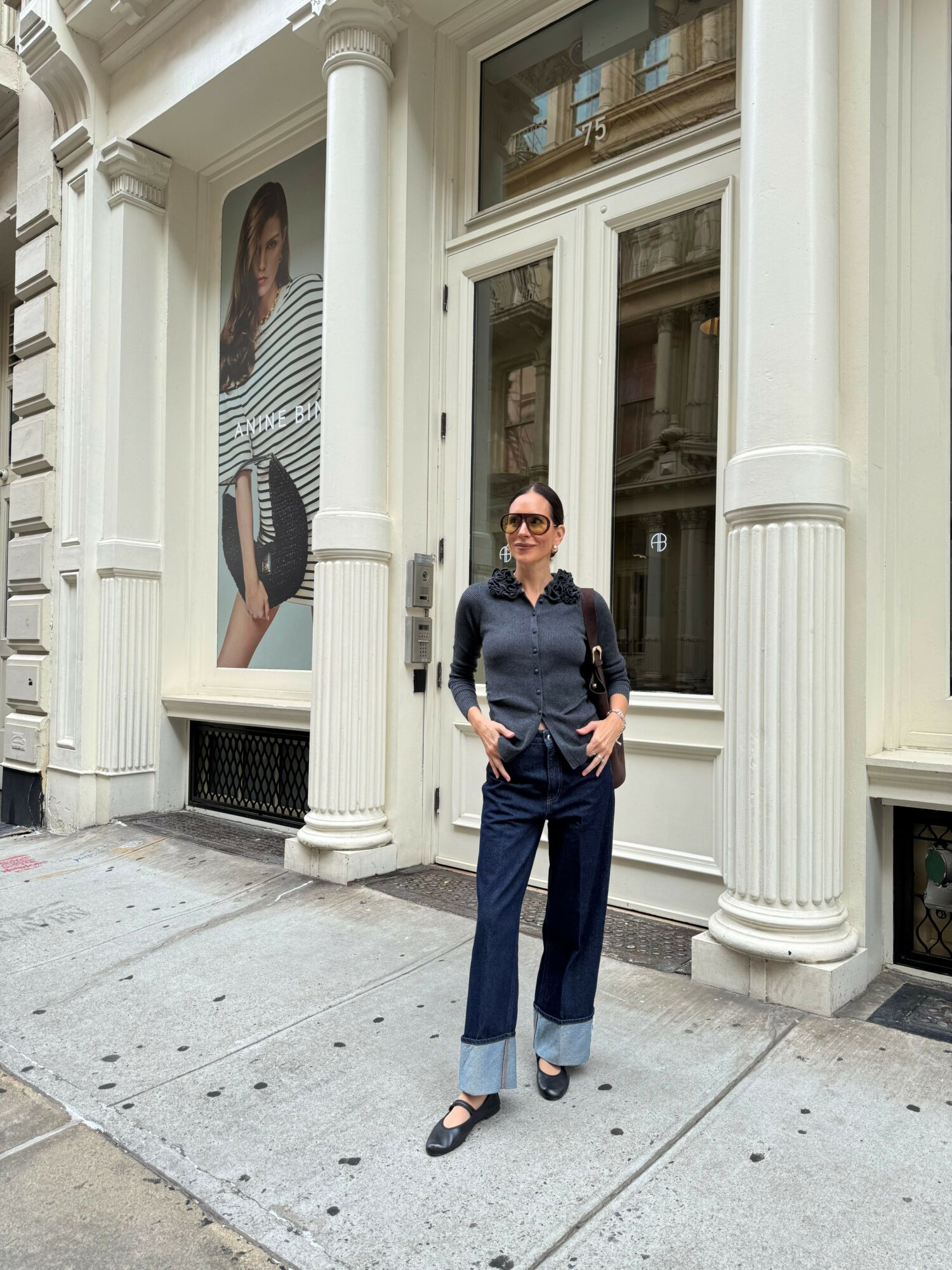 Person standing on sidewalk in front of building with large window and white columns, holding a phone, wearing dark clothing.