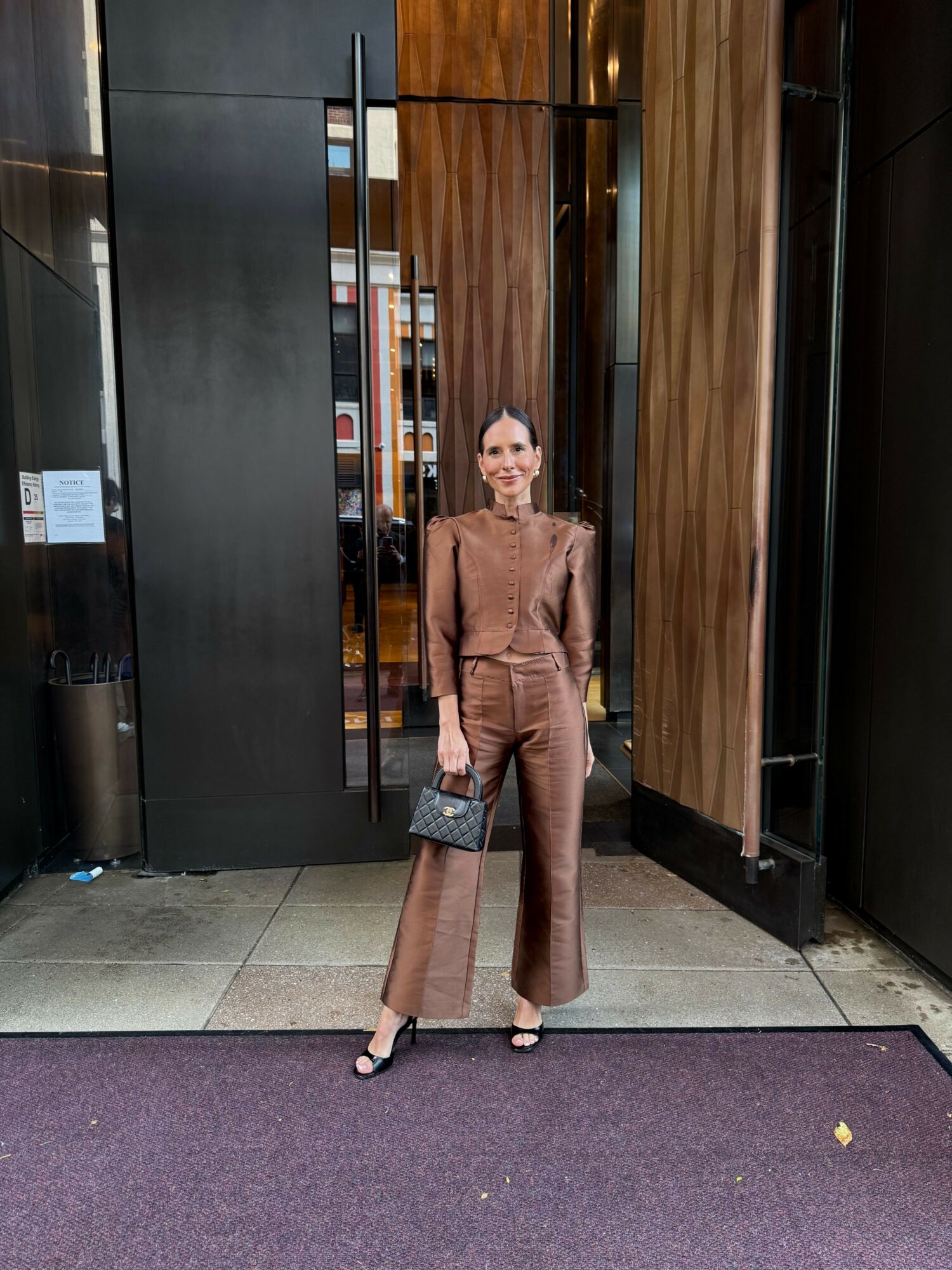 Woman in brown suit standing on purple carpet outside building with wooden and black wall background.