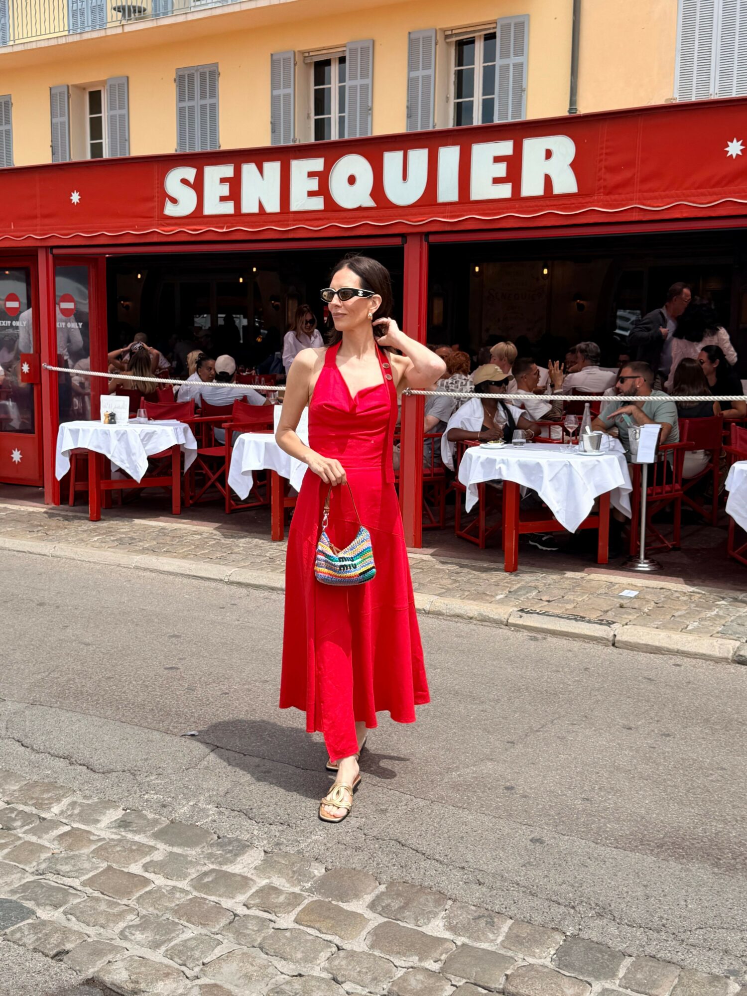 Woman in red dress walking on cobblestone street outside restaurant with outdoor seating.