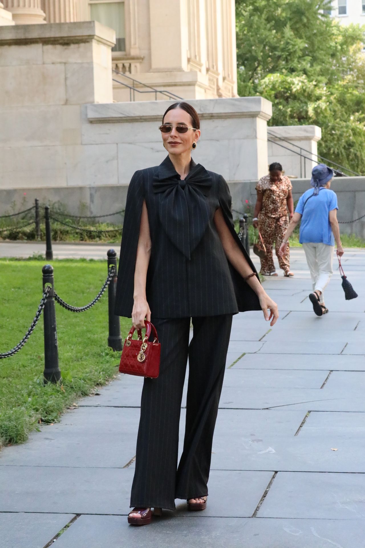 Woman in black outfit holding a red bag standing outdoors near a building with greenery in the background.