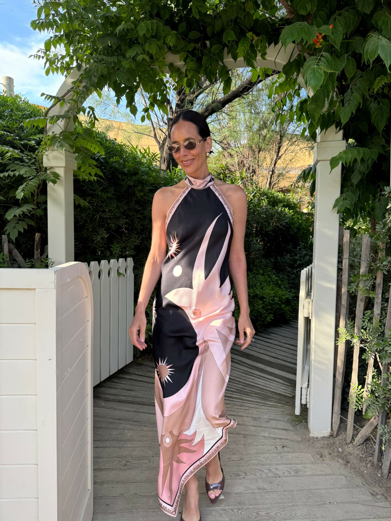 Woman in a black and pink dress standing outdoors on a wooden deck, surrounded by greenery and trees.