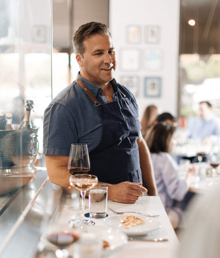 Man in a blue shirt and apron standing at a bar with wine glasses and plates, smiling, in a bright restaurant.
