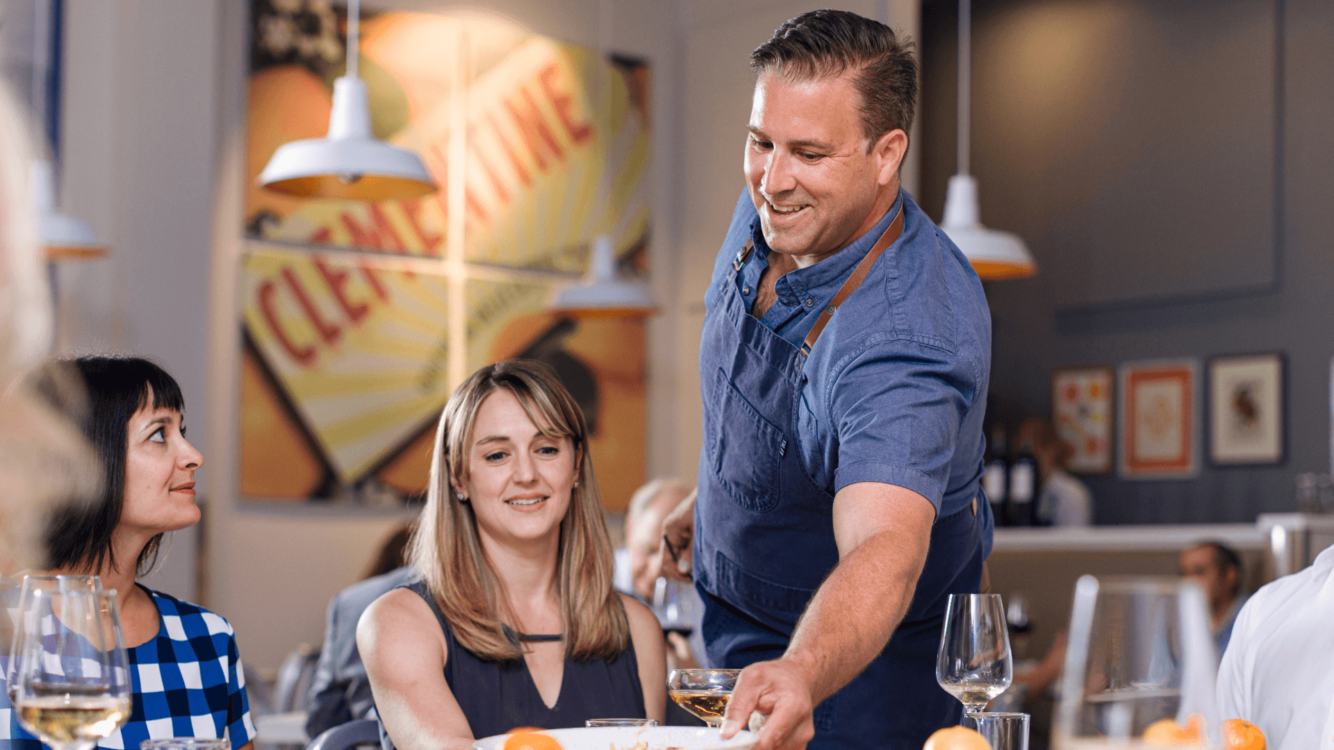 Man serving food to two women at a restaurant table, smiling, with a colorful background and glasses on the table.