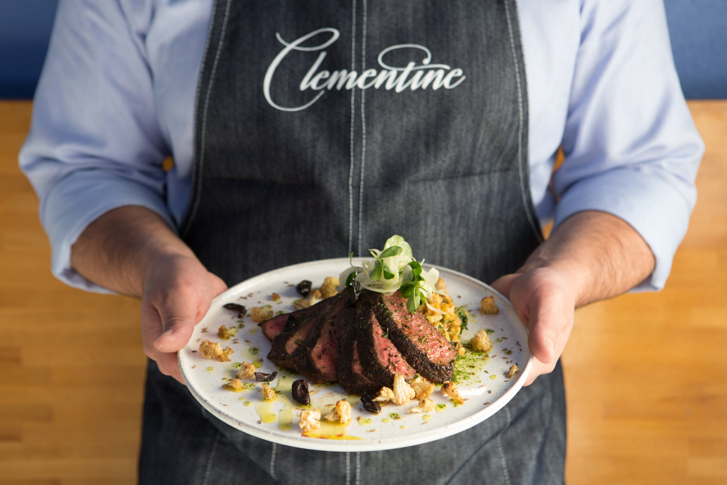 Person holding a plate with sliced steak, garnished with vegetables and sauce, wearing an apron with 'Clementine' written on it.