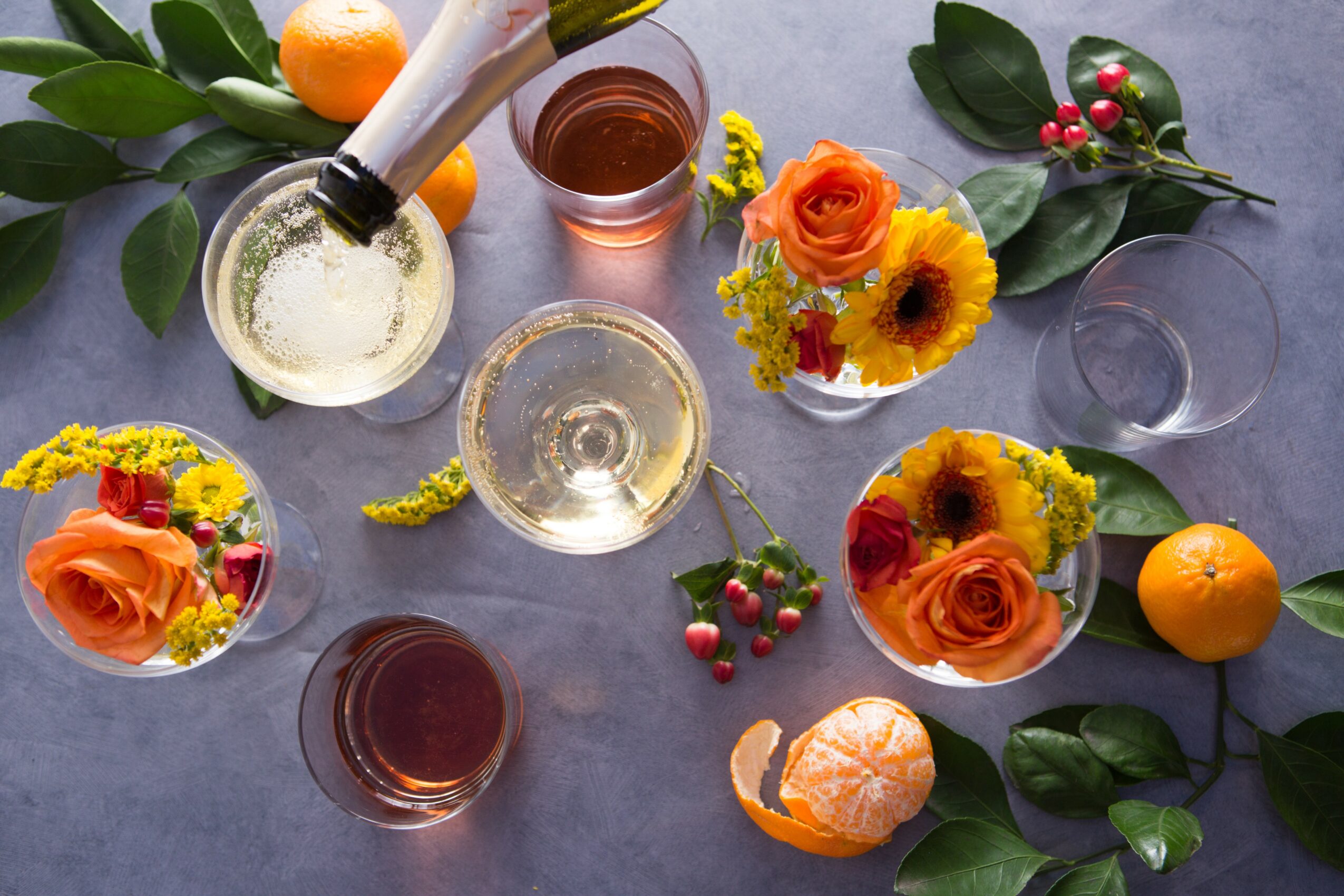 Top-down view of glasses with drinks, flowers, oranges, and berries on a gray surface.