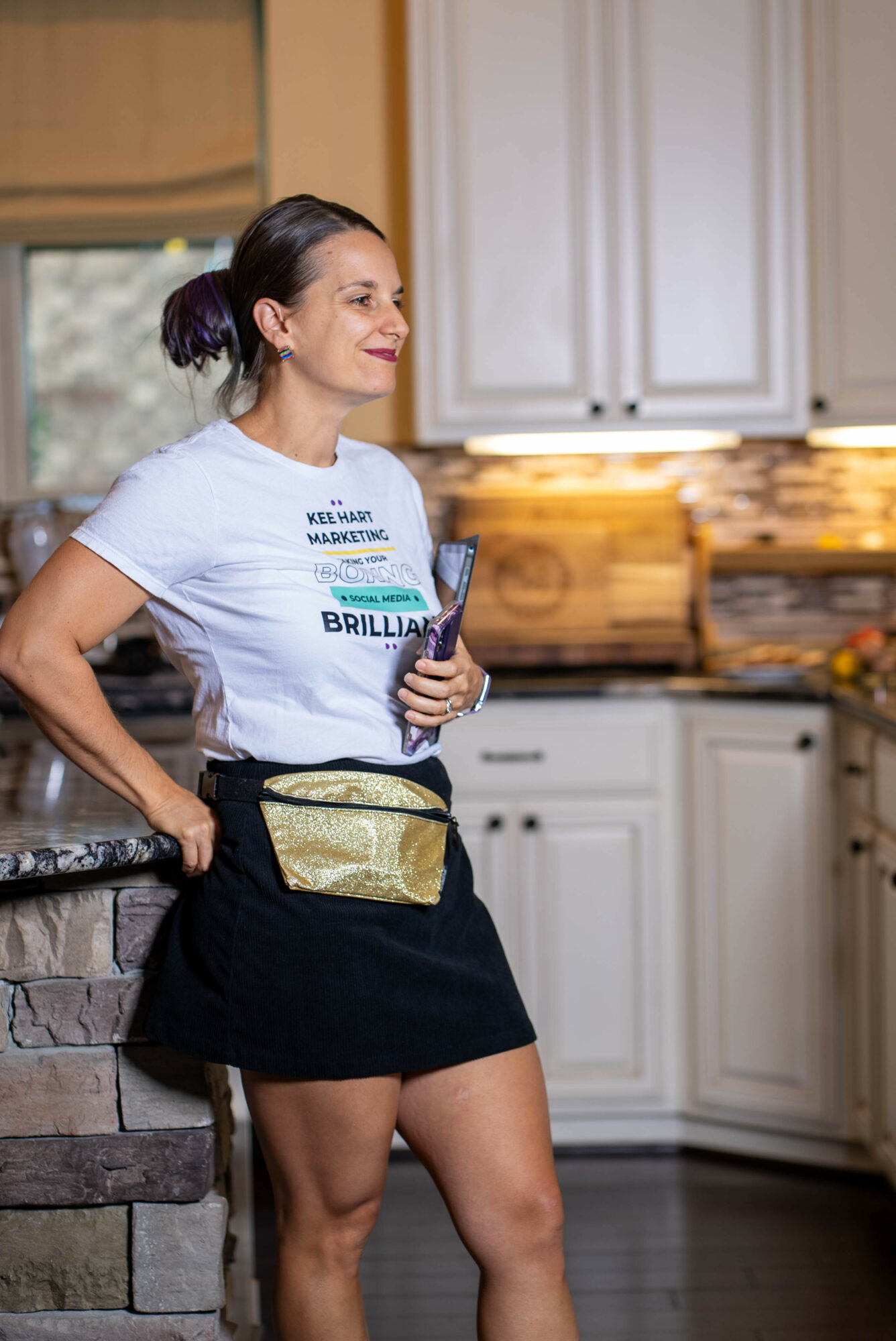 Woman standing in a kitchen, holding a glass, smiling, wearing a white t-shirt and black skirt, with a gold fanny pack.