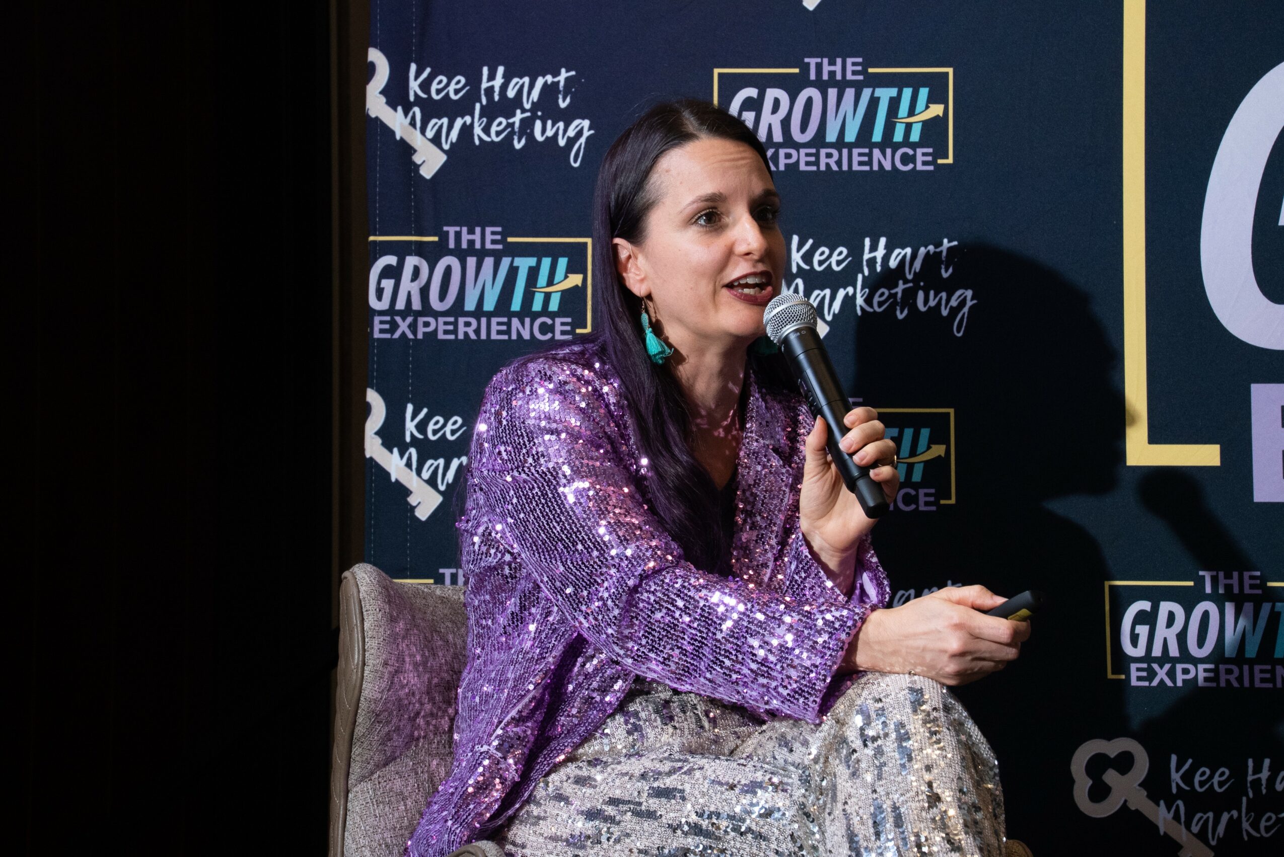 Woman speaking into a microphone at a conference, with a dark background and event branding behind her.
