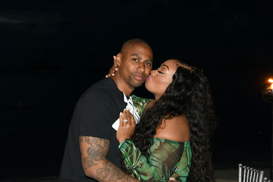 A man and woman pose closely at night, the woman kisses the man's cheek, both looking at the camera, dark background.
