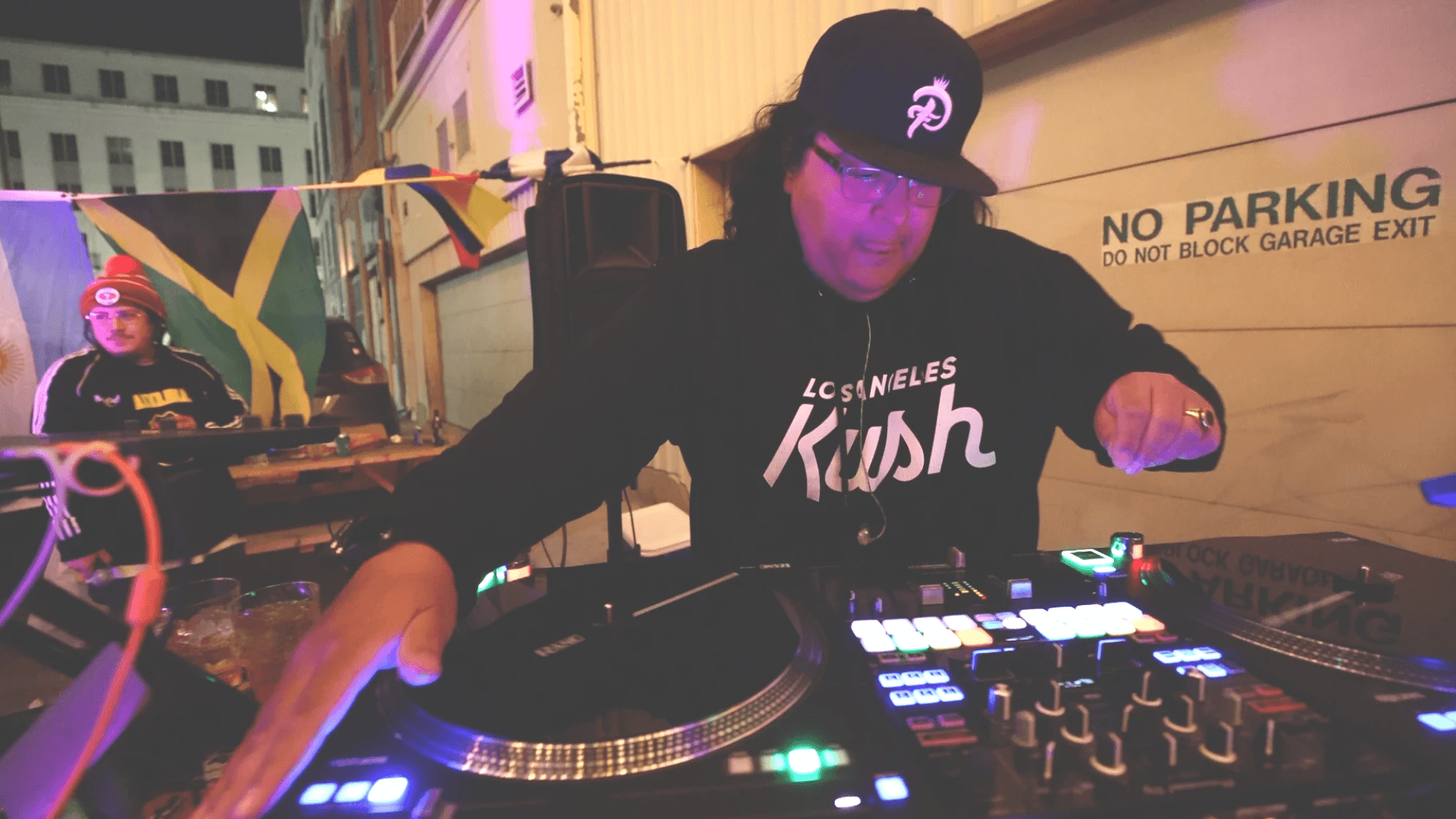 DJ wearing a cap and hoodie, operating DJ equipment at night, with a person in the background near a building with a Jamaican flag.