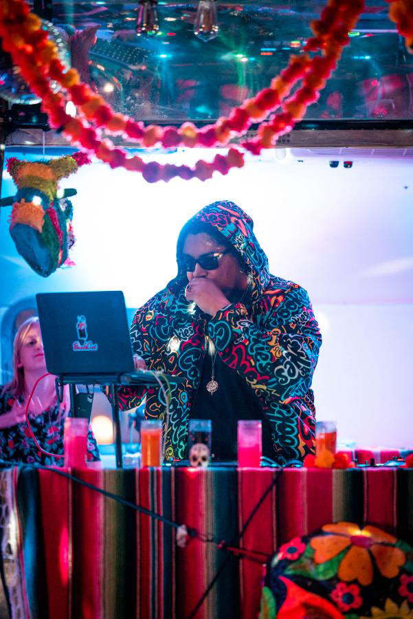 Person wearing colorful clothing and sunglasses, DJing at a decorated party with balloons and vibrant decorations.