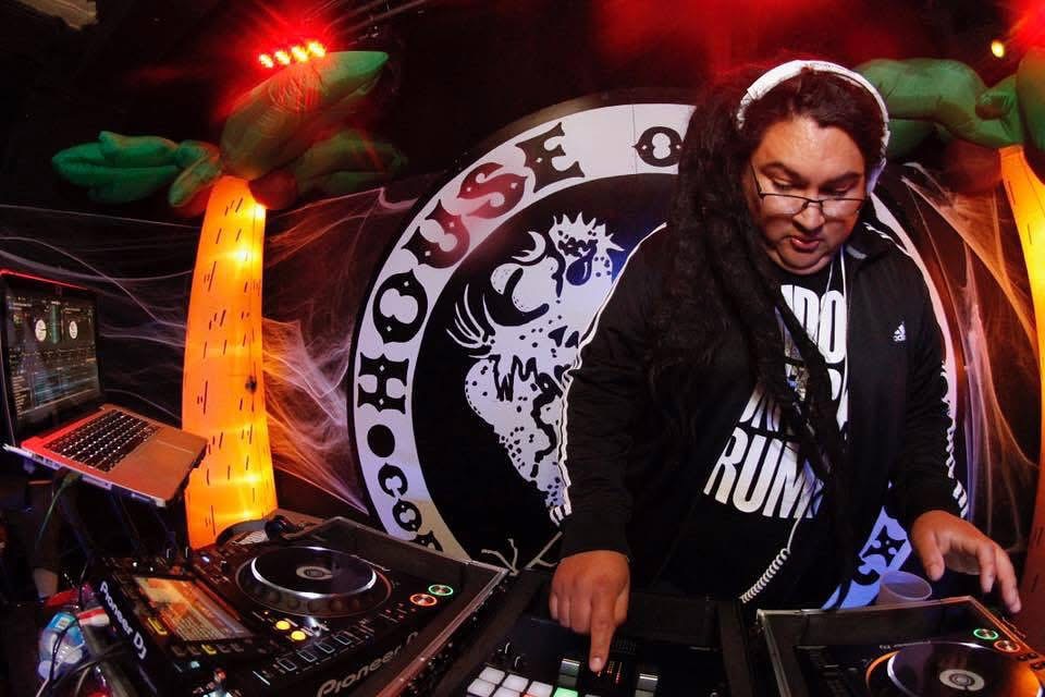 Female DJ with glasses and curly hair operating DJ equipment at a club with a logo in the background.