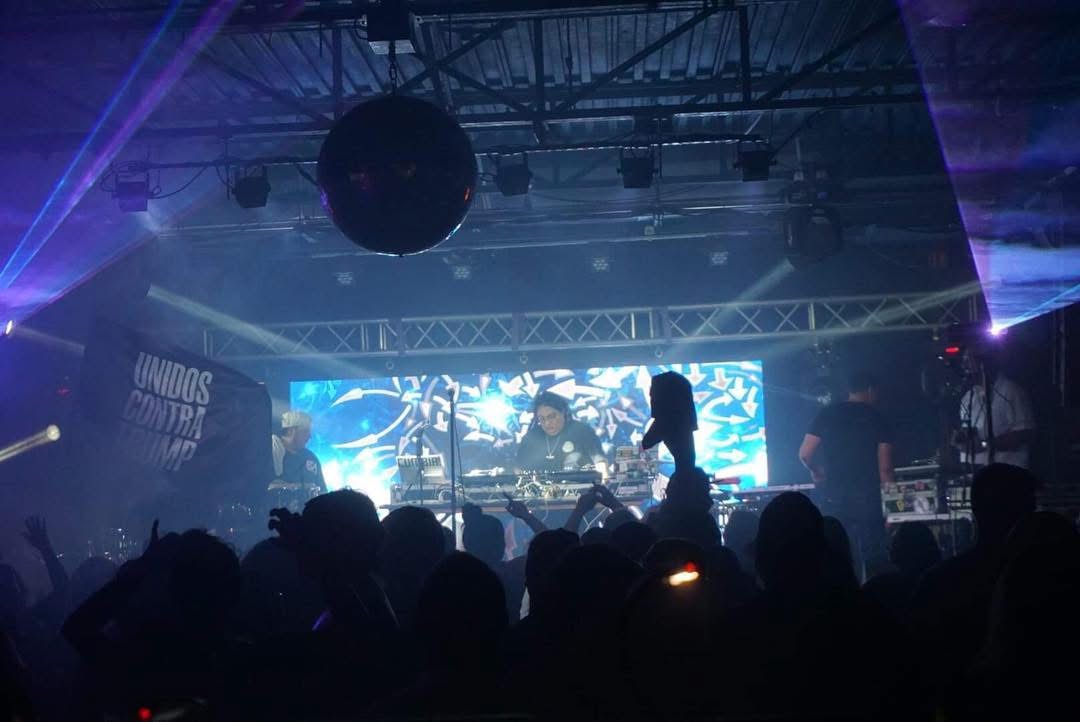 Concert scene with stage, performers, and audience silhouettes, illuminated by colorful lights and screens in the background.