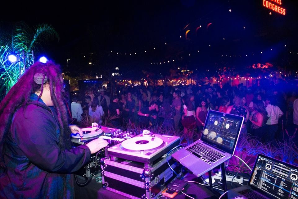 Person with long hair and headphones DJing at a night event with a crowd and colorful lights.