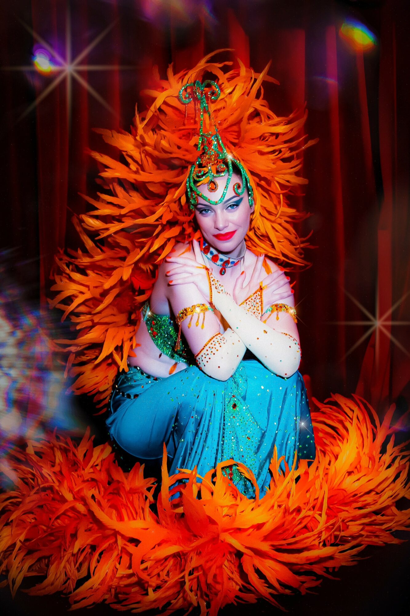 Performer in colorful costume with large orange feather headdress, sitting on stage with red curtains behind.