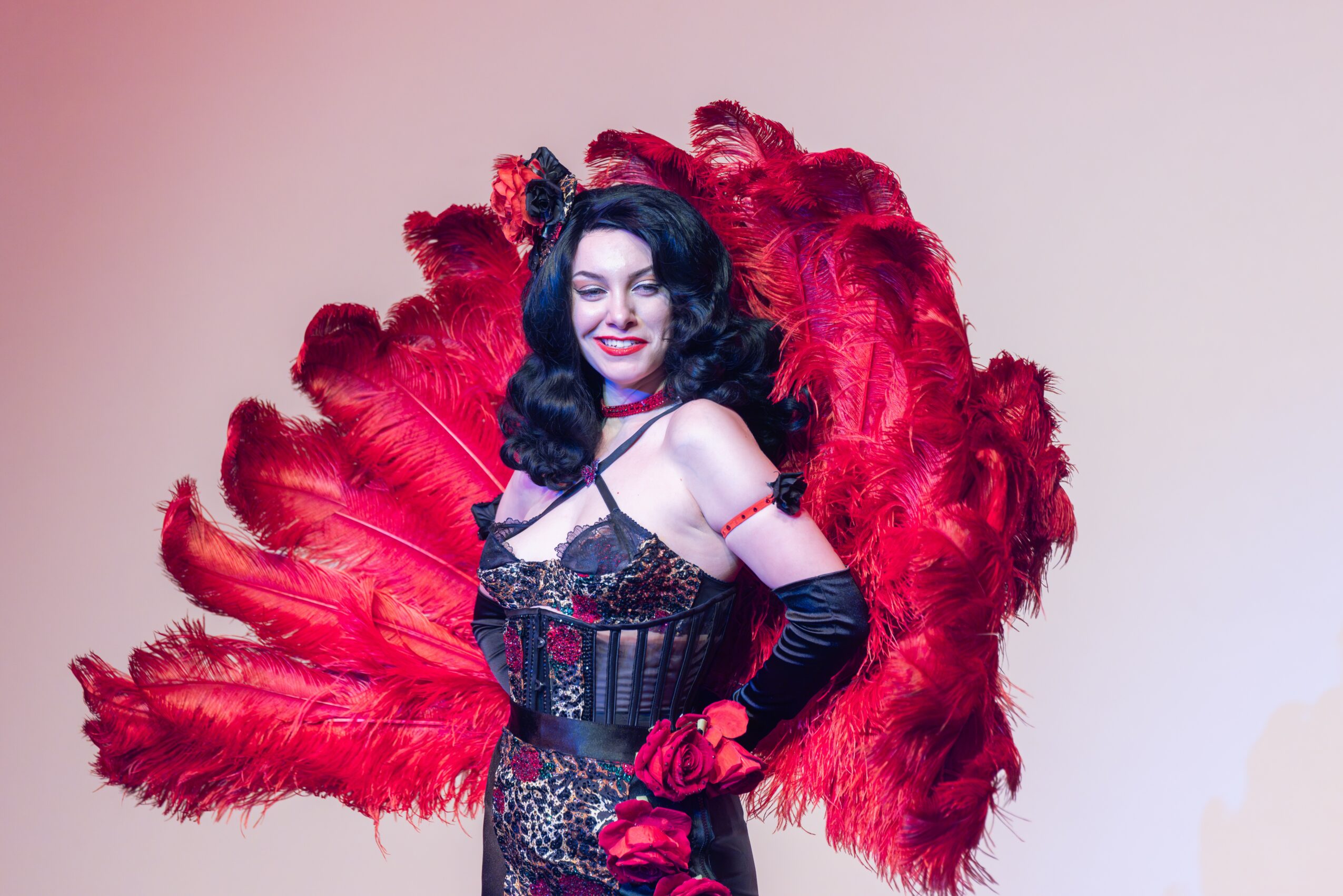 Woman in a black lace dress with red feathered wings, smiling, against a plain background.