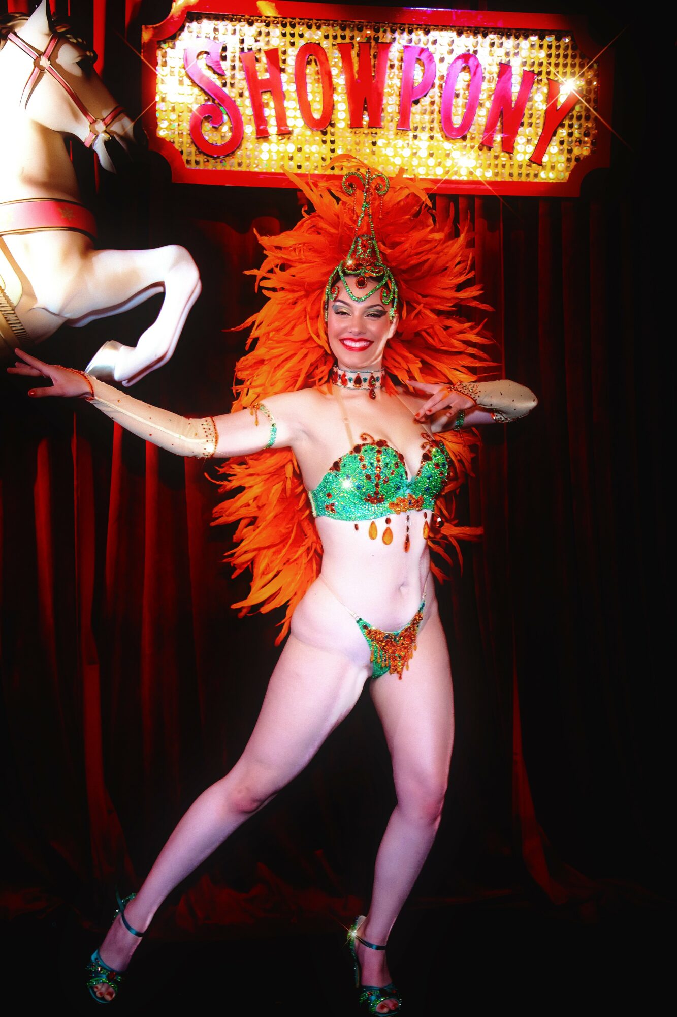 Performer in colorful costume with large orange feathered headdress on stage with a sign reading 'SHOW PONY'.