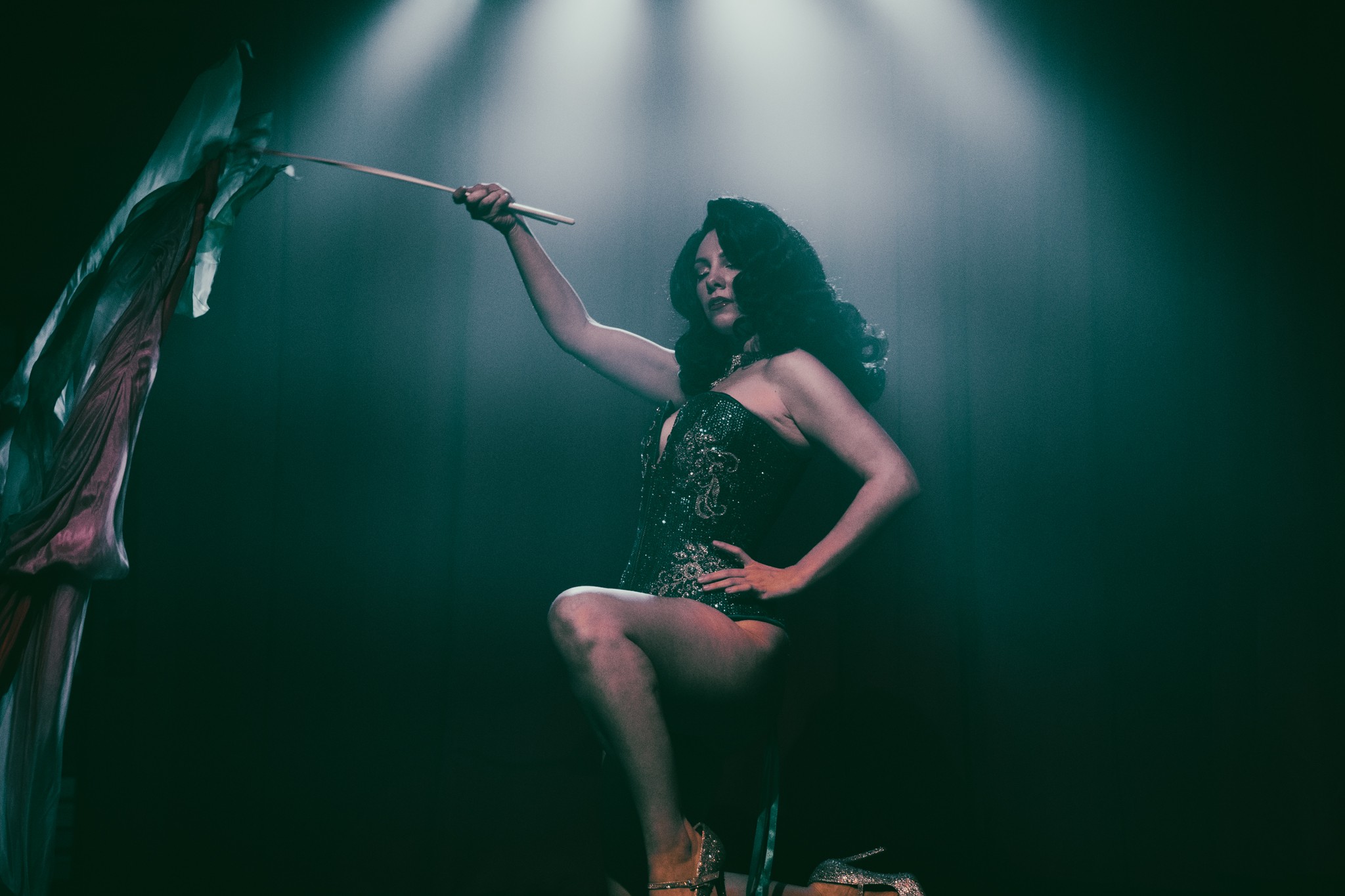 Woman with curly hair in a black dress holding a whip, sitting on a stool in a dimly lit room.