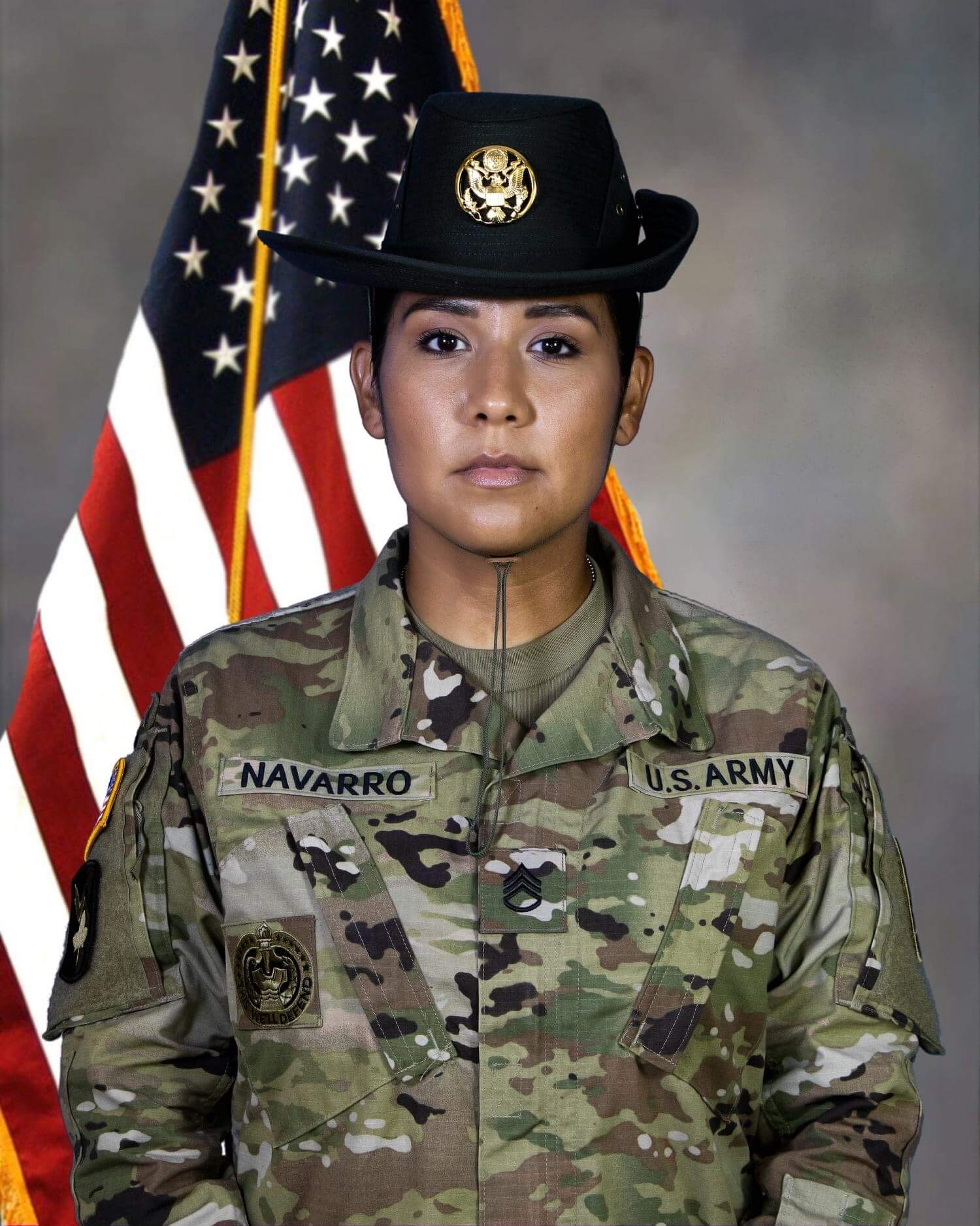 Female soldier in U.S. Army uniform with a hat, standing in front of an American flag, looking serious.