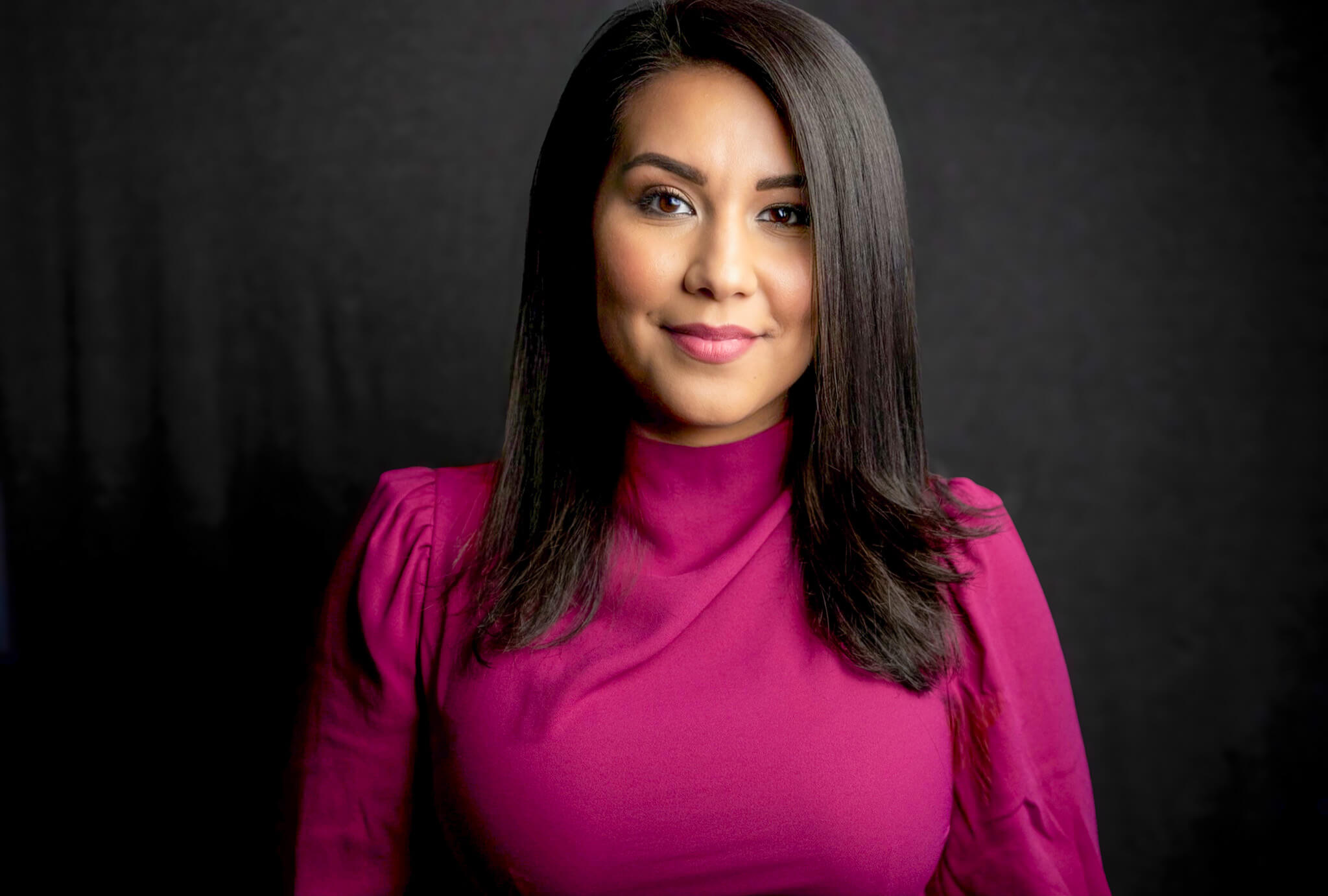 Woman with shoulder-length dark hair wearing a magenta top, smiling against a dark background.