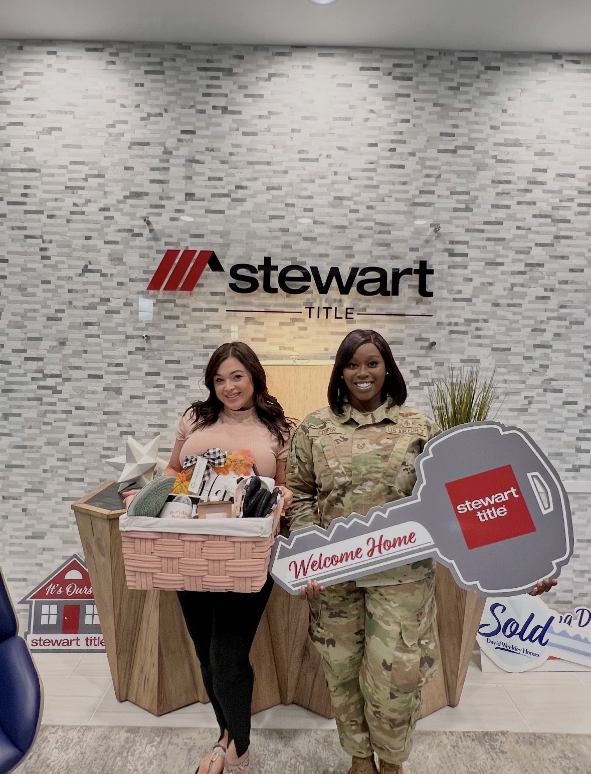 Two women stand in front of a Stewart Title display, holding a large key and a welcome sign, smiling at the camera.