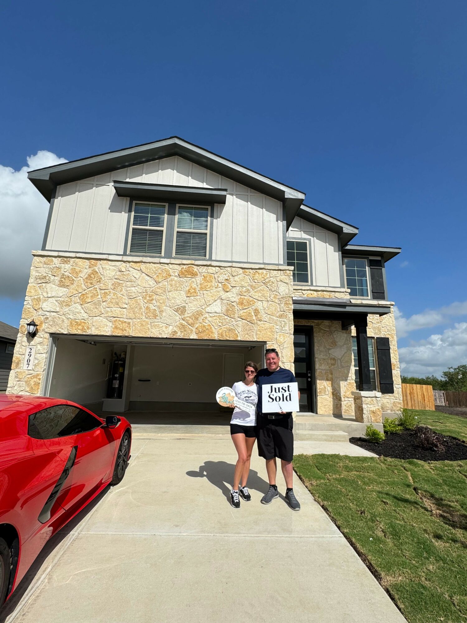 Two children stand on a sidewalk in front of a modern house with a stone facade, holding signs, with a red car nearby.