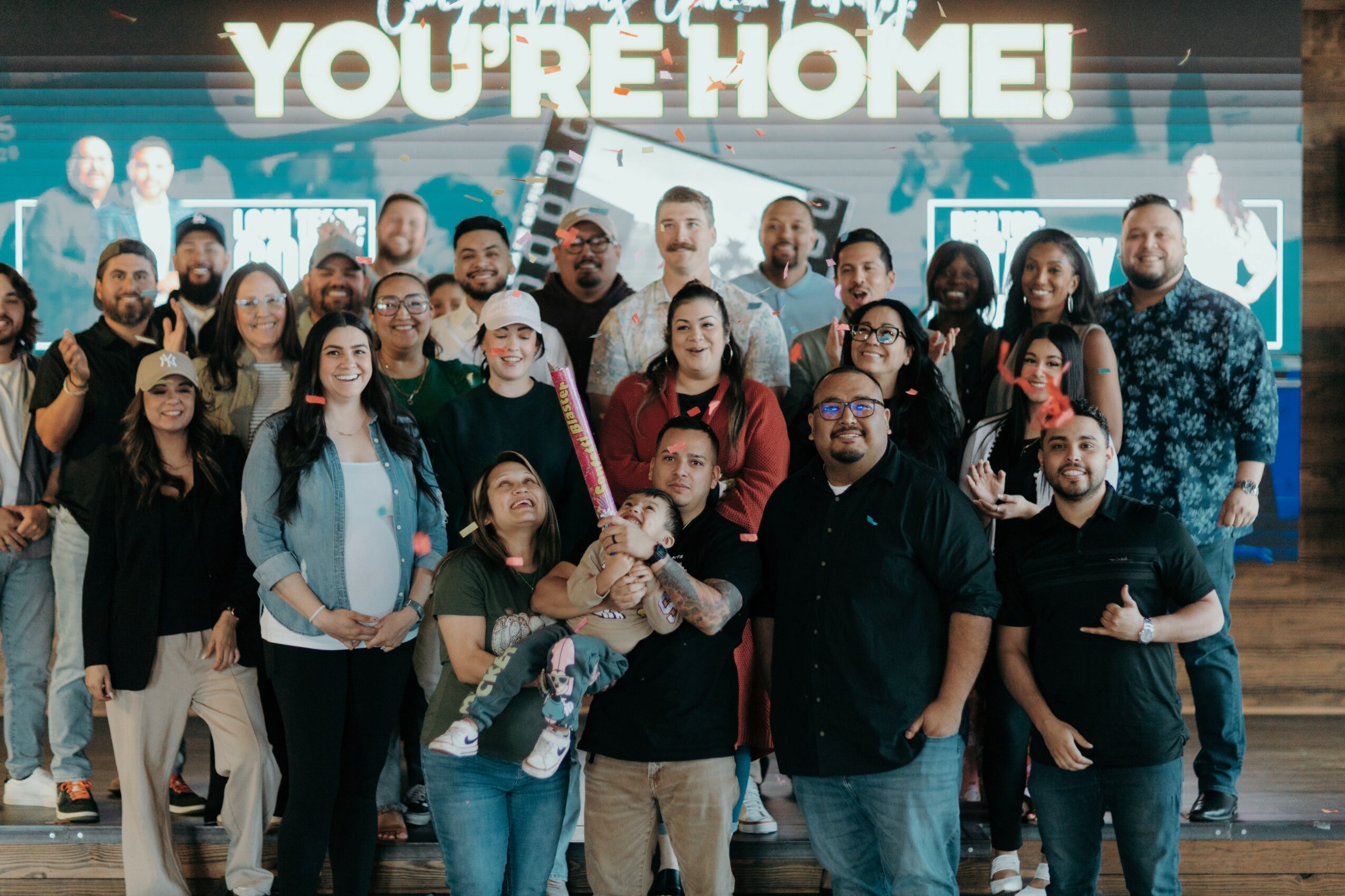 Group of diverse people smiling and holding a sign that says 'You're Home!' inside a building.