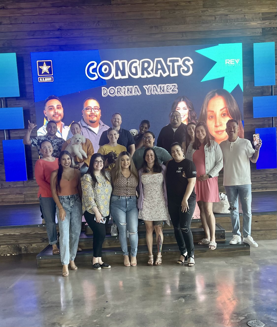 Group of people standing on stage in front of a congratulatory screen with the name Dorina Yanez, some smiling and posing.