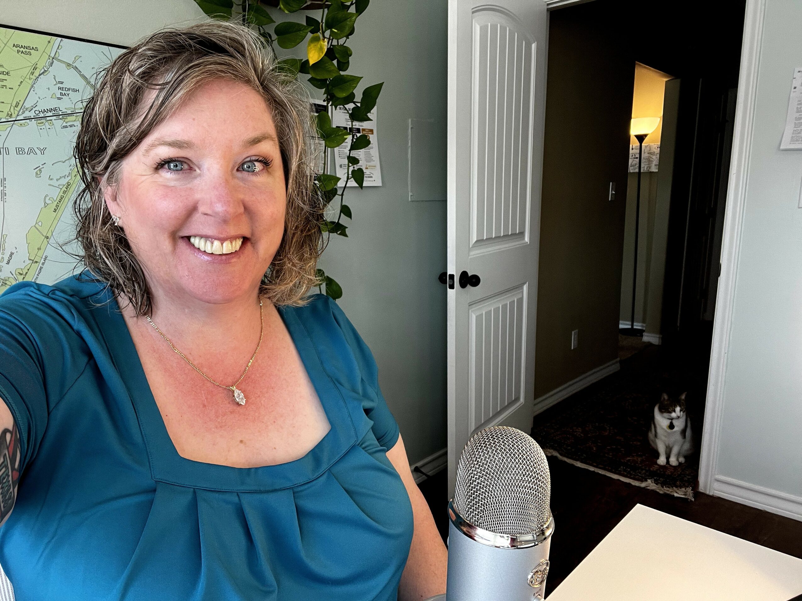 Smiling woman with short hair in a blue top taking a selfie, with a cat in the background near a doorway.
