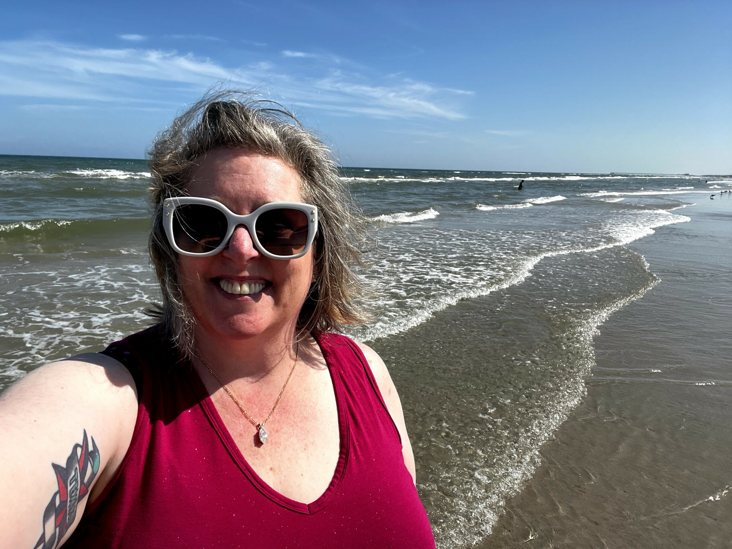 Woman with sunglasses smiling at the beach, ocean waves in background, wearing a red top, sunny day.