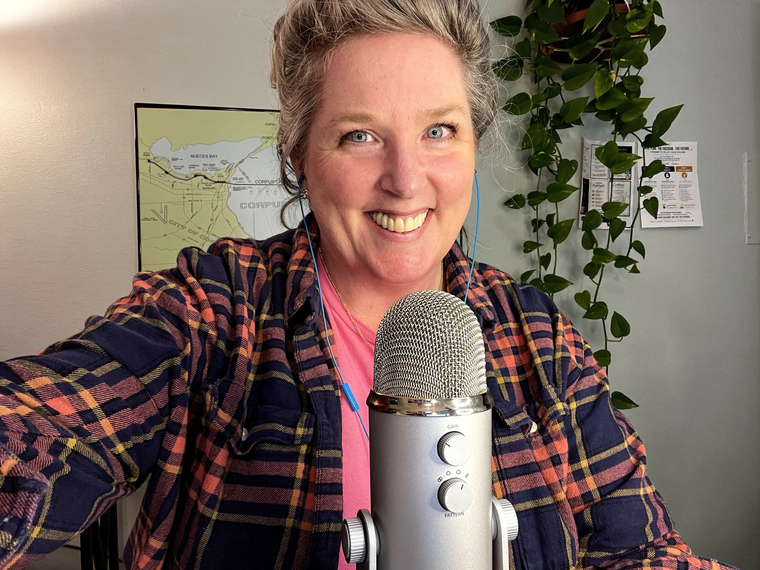 Smiling woman with short gray hair taking a selfie in front of a microphone, with a plant and map in background.