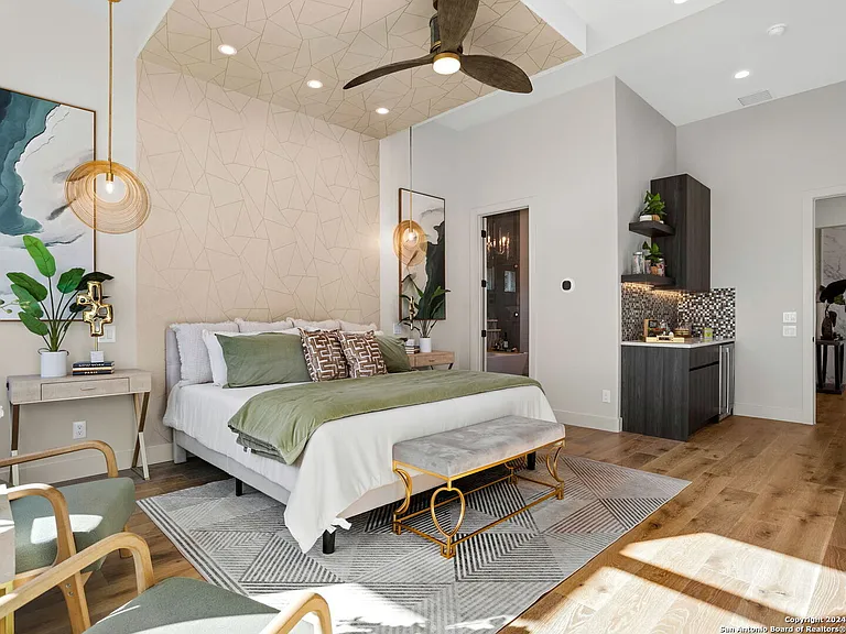 Modern bedroom with bed, nightstands, plants, and a ceiling fan, with a kitchen area visible in the background.