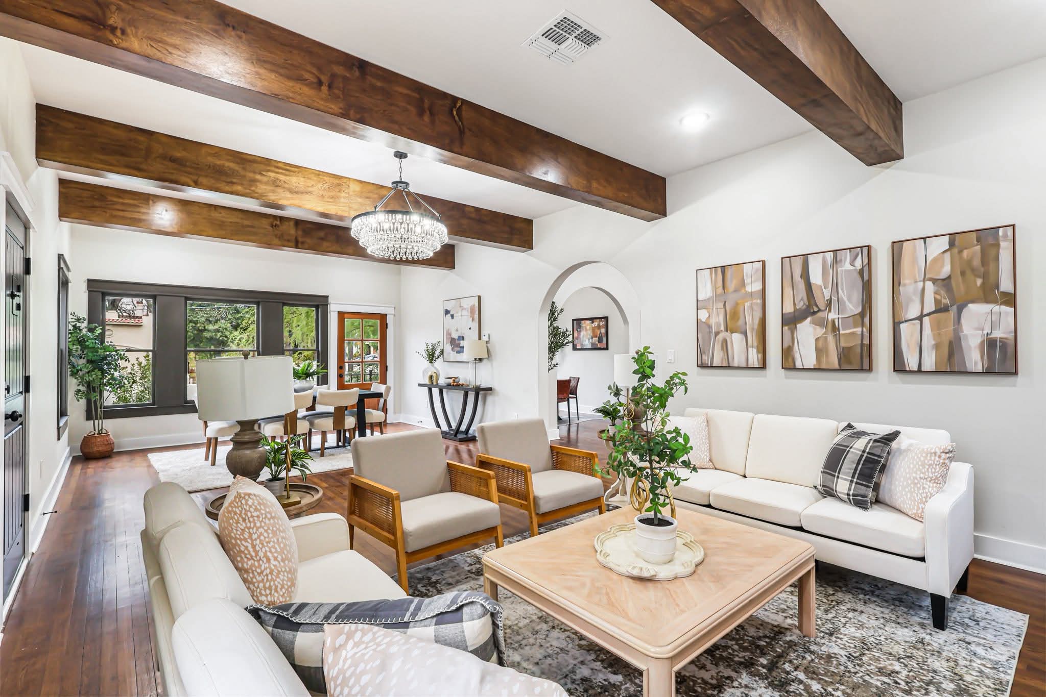 Living room with white sofa, wooden chairs, large windows, wooden beams, and artwork on wall.