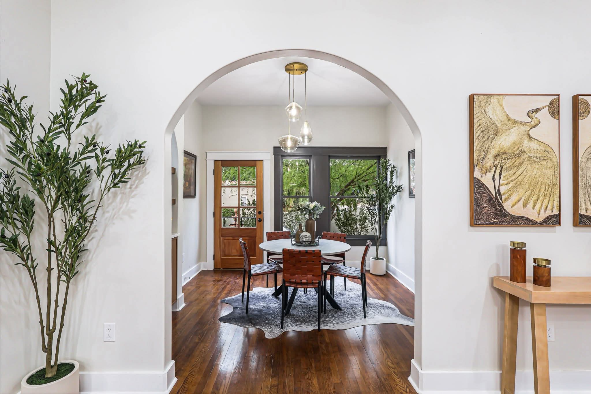 Dining room with round table, four chairs, and a chandelier, viewed through an arched doorway, with a door and window in background.