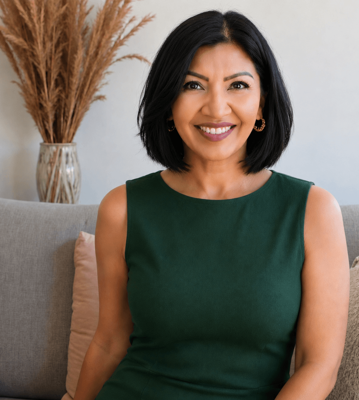 Woman with shoulder-length black hair smiling, wearing a green sleeveless top, sitting on a sofa with a beige cushion, in a room with a vase of dried plants in the background.