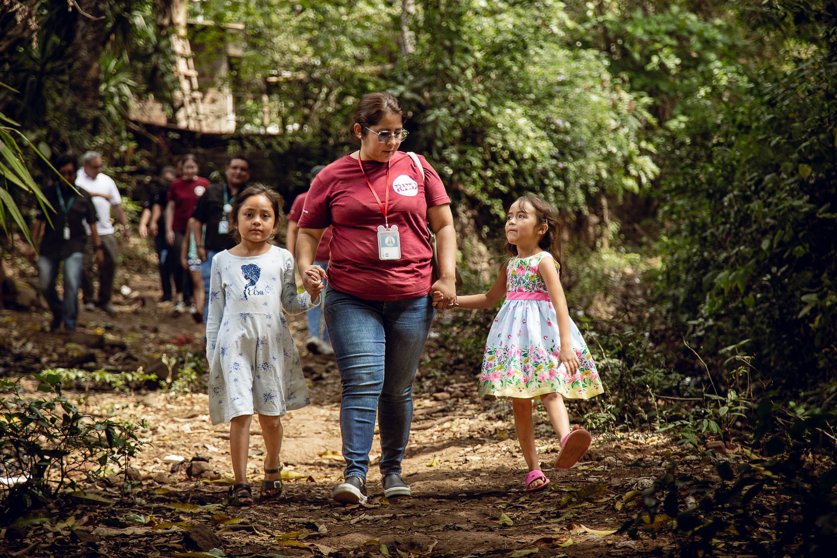 Woman walking with two young girls outdoors in a wooded area, holding hands, with other people in the background.