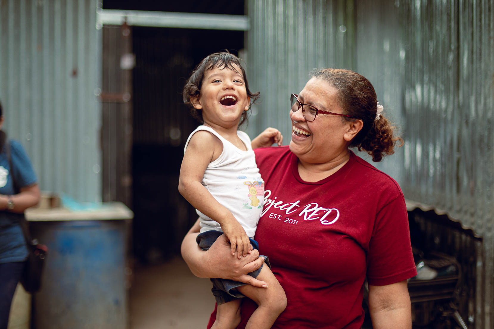 Woman holding a laughing child outdoors, both smiling, in front of a corrugated metal wall.