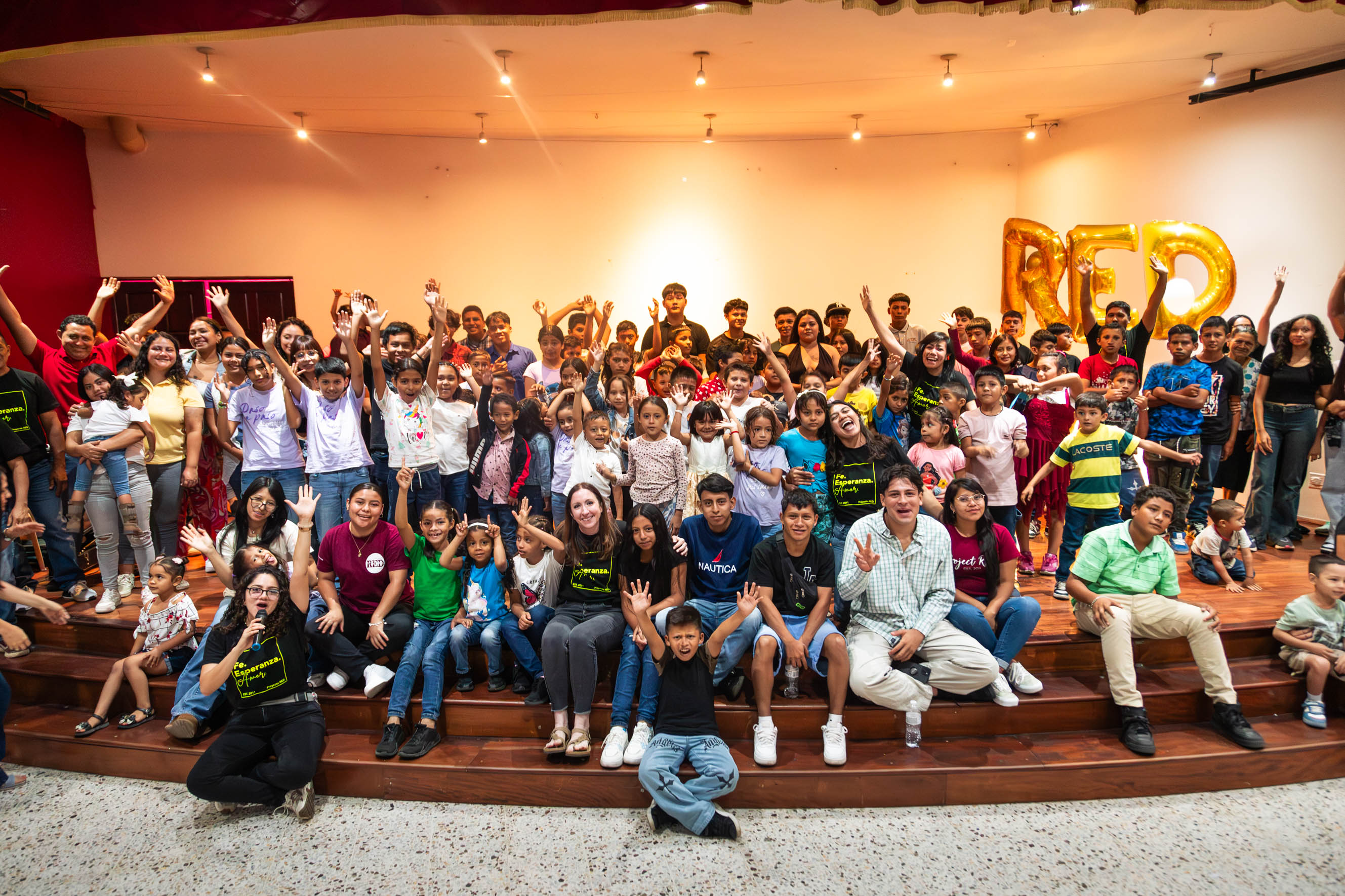 Large group of children and adults on stage, some waving, in a spacious auditorium with warm lighting.
