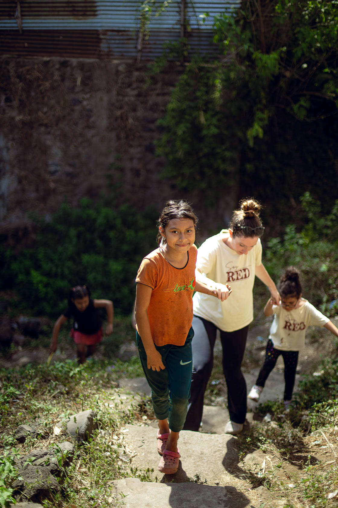 Four children walking up a dirt path outdoors, with greenery and a brick wall in the background.
