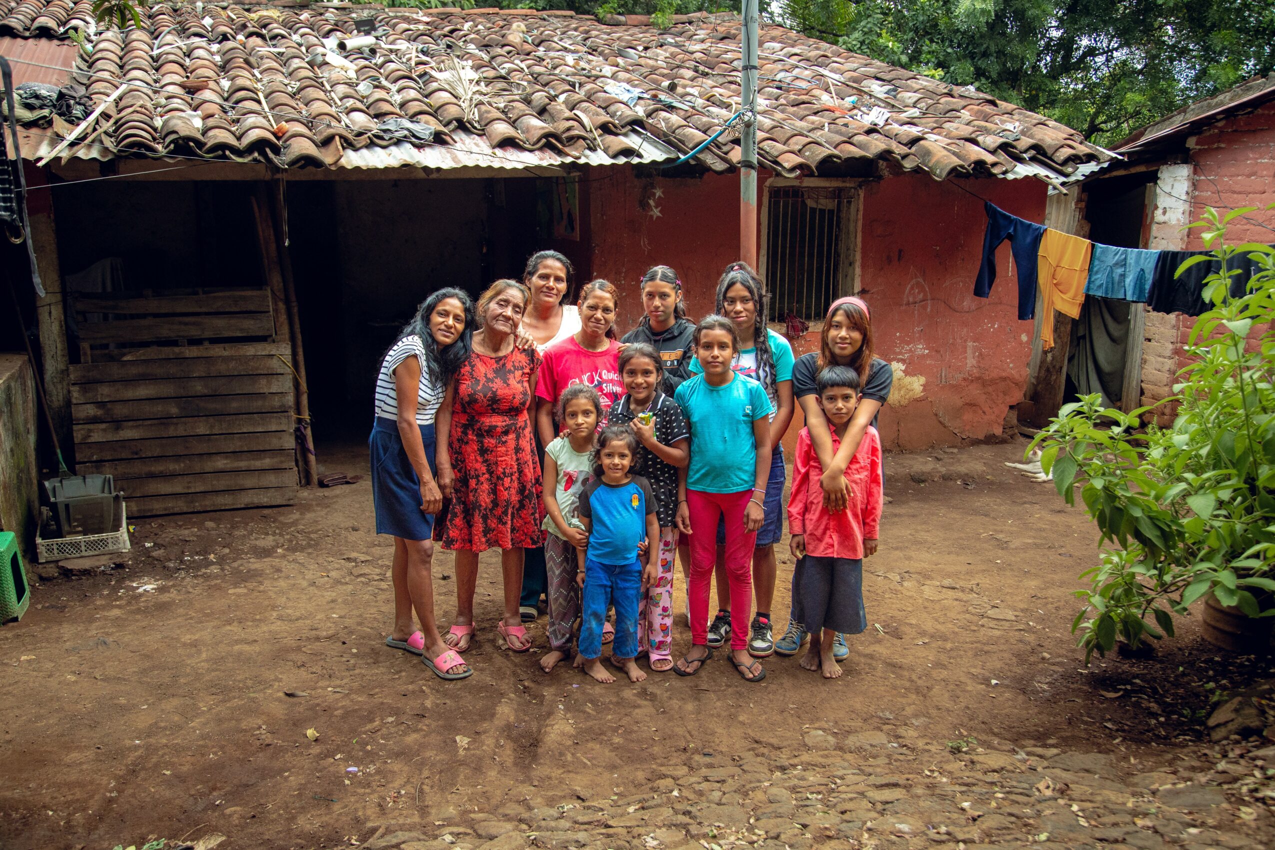 Group of children and adults standing outdoors in front of rustic buildings, some wearing colorful clothing, with laundry hanging on a line.