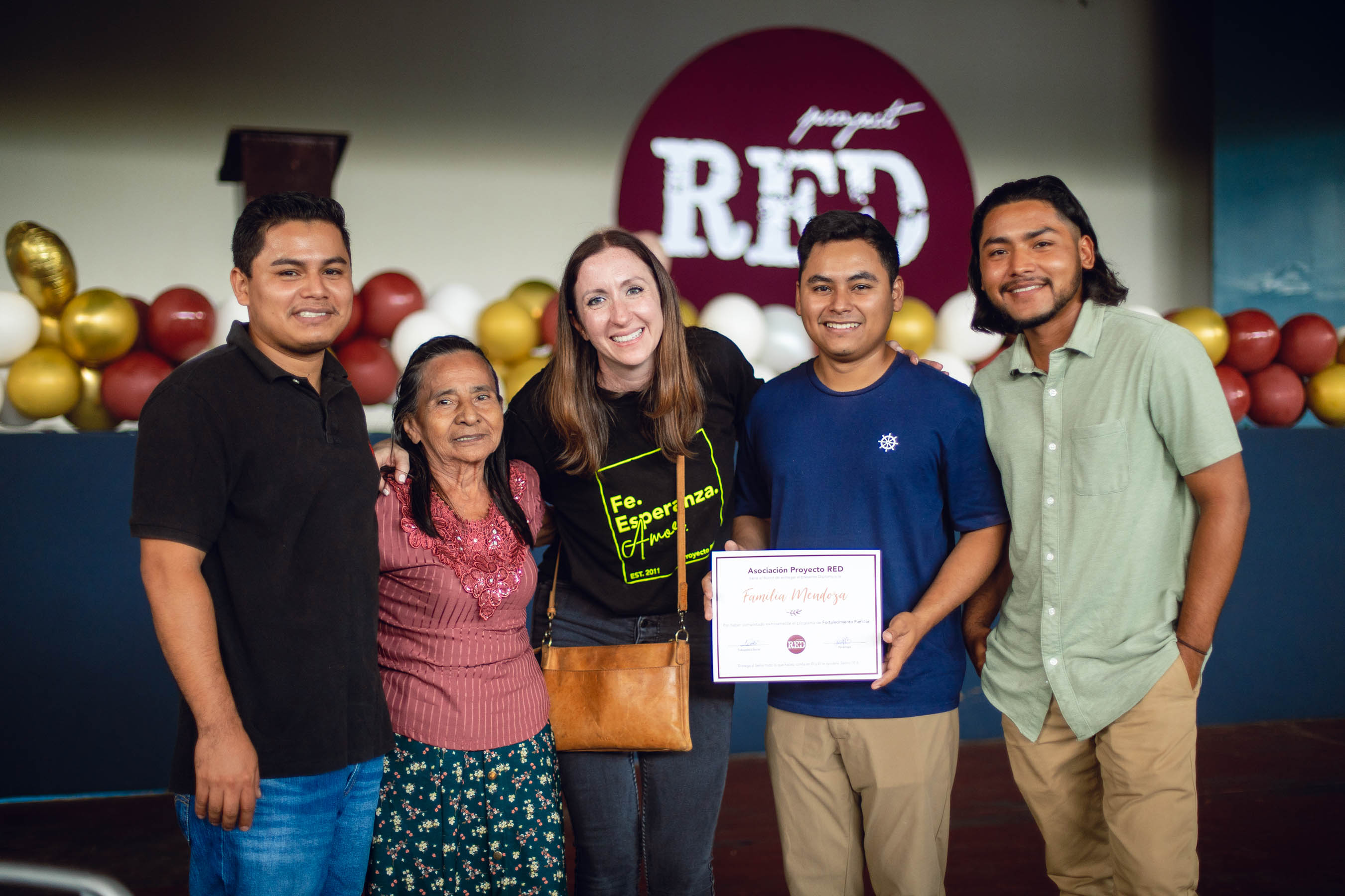 Group of five people smiling, one holding a certificate, in front of a backdrop with balloons and a sign that says 'RED'.