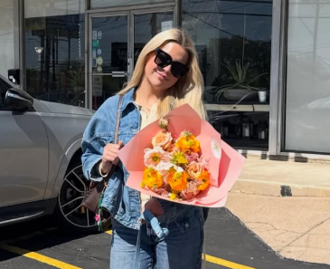 Woman with blonde hair wearing sunglasses and denim jacket holding a bouquet of flowers outside a building.