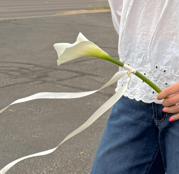 Person holding a white flower with long ribbon, standing on a paved surface, wearing a white top and blue jeans.