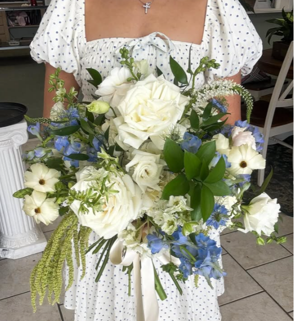 Person holding a large bouquet of white and blue flowers with green leaves, wearing a white polka dot dress and a cross necklace.
