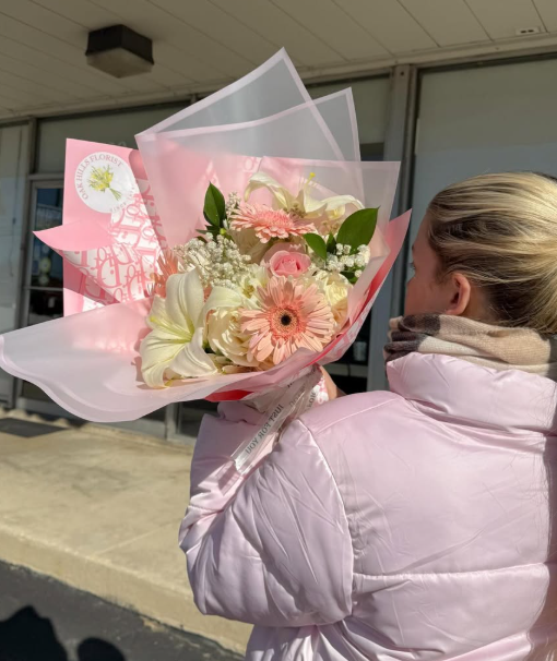 Person holding a bouquet of pink and white flowers, wearing a light pink jacket, with a building in the background.