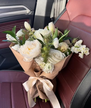 Bouquet of white flowers with green leaves wrapped in brown paper and tied with a beige ribbon, placed on a car seat.