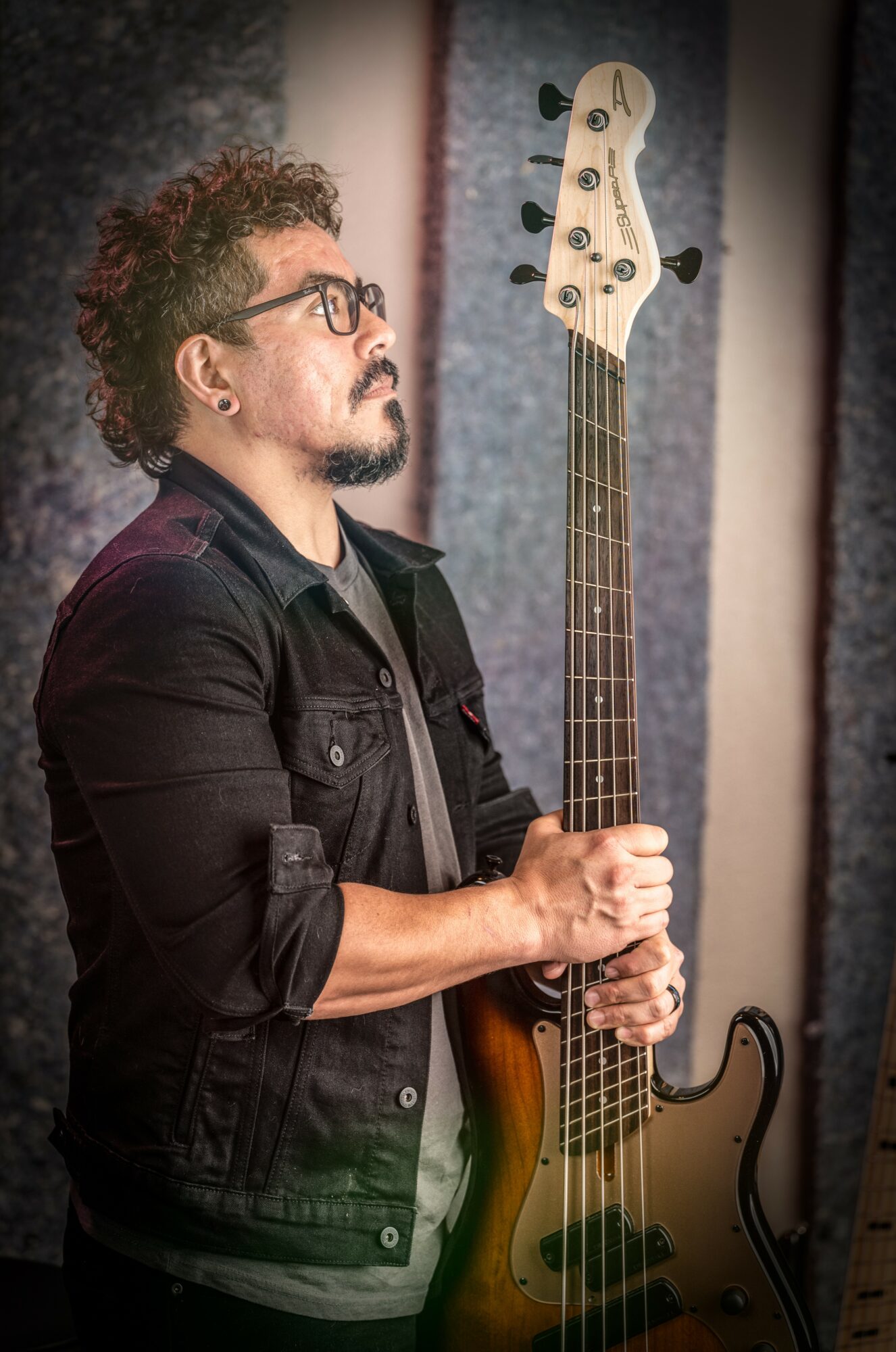 Man with curly hair and glasses holding a bass guitar, wearing a black jacket, in a recording studio.