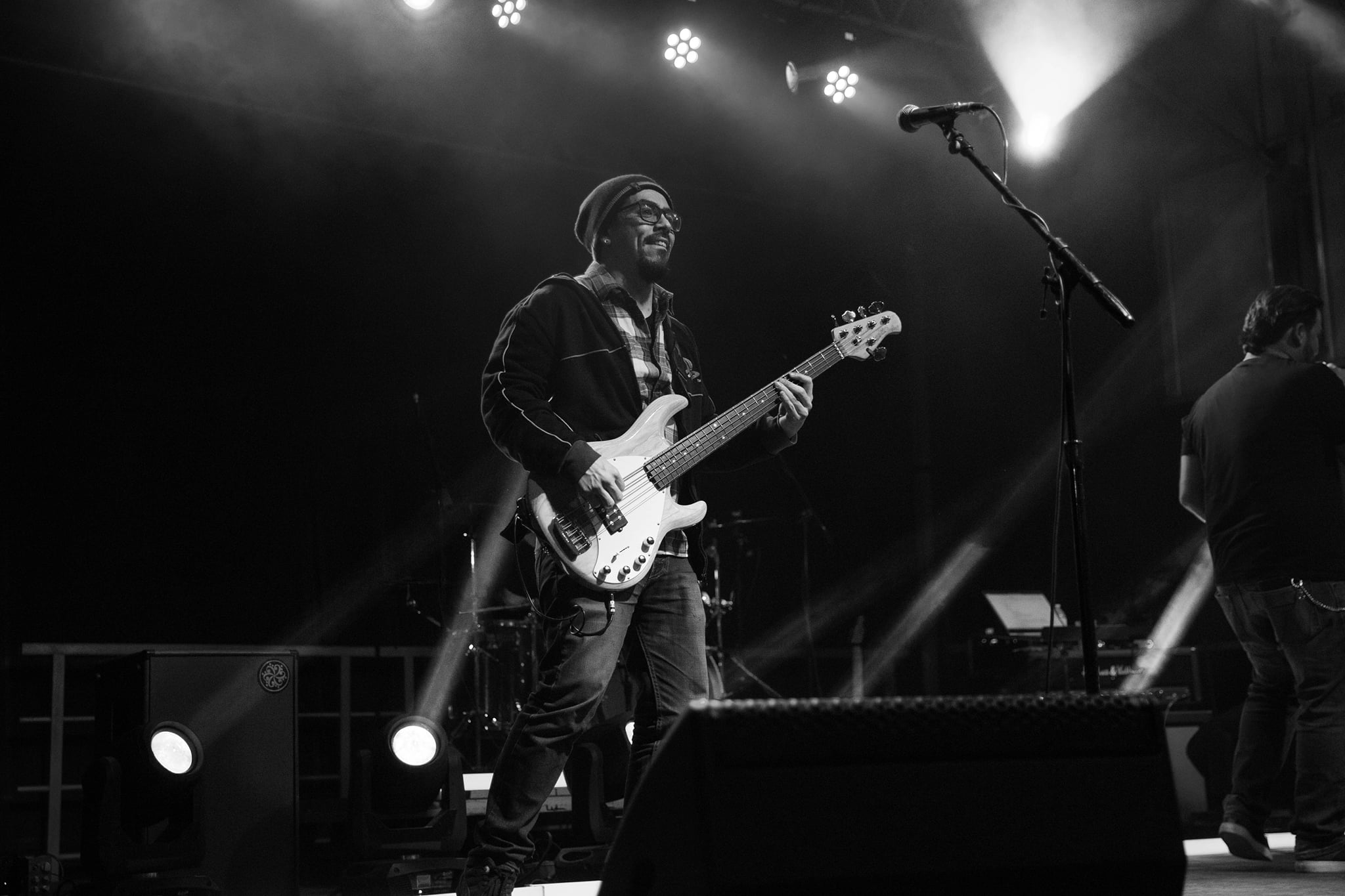 Musician playing bass guitar on stage with bright lights and fog, wearing glasses and a hat.
