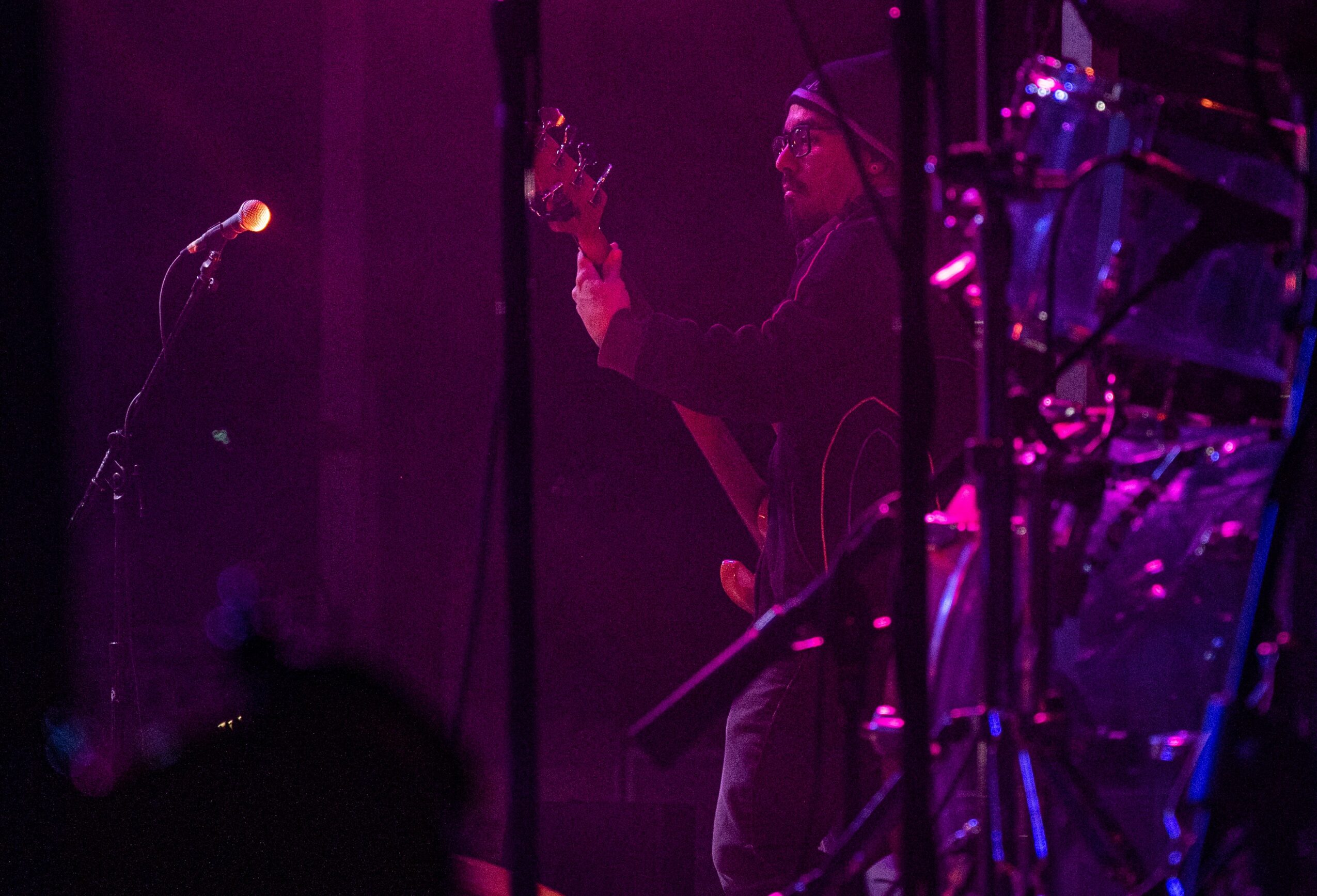 Person on stage holding a guitar, with musical equipment and a microphone, illuminated by purple lighting.